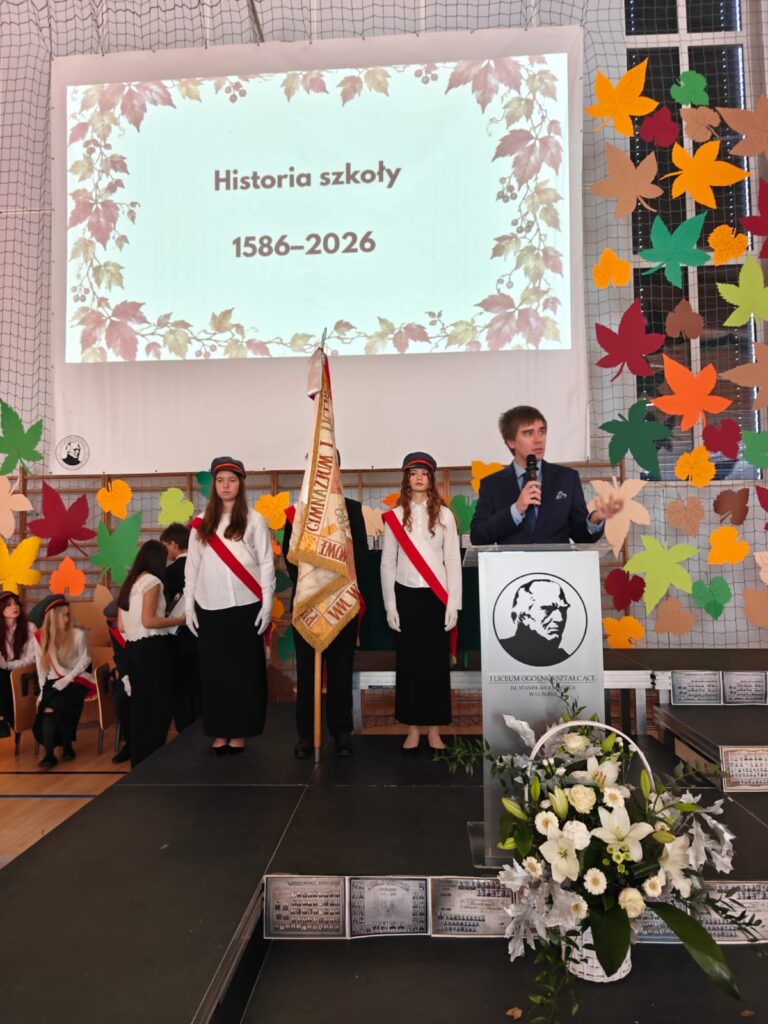 Poczet sztandarowy stoi przed ekranem z napisem „Historia szkoły 1586–2026”.