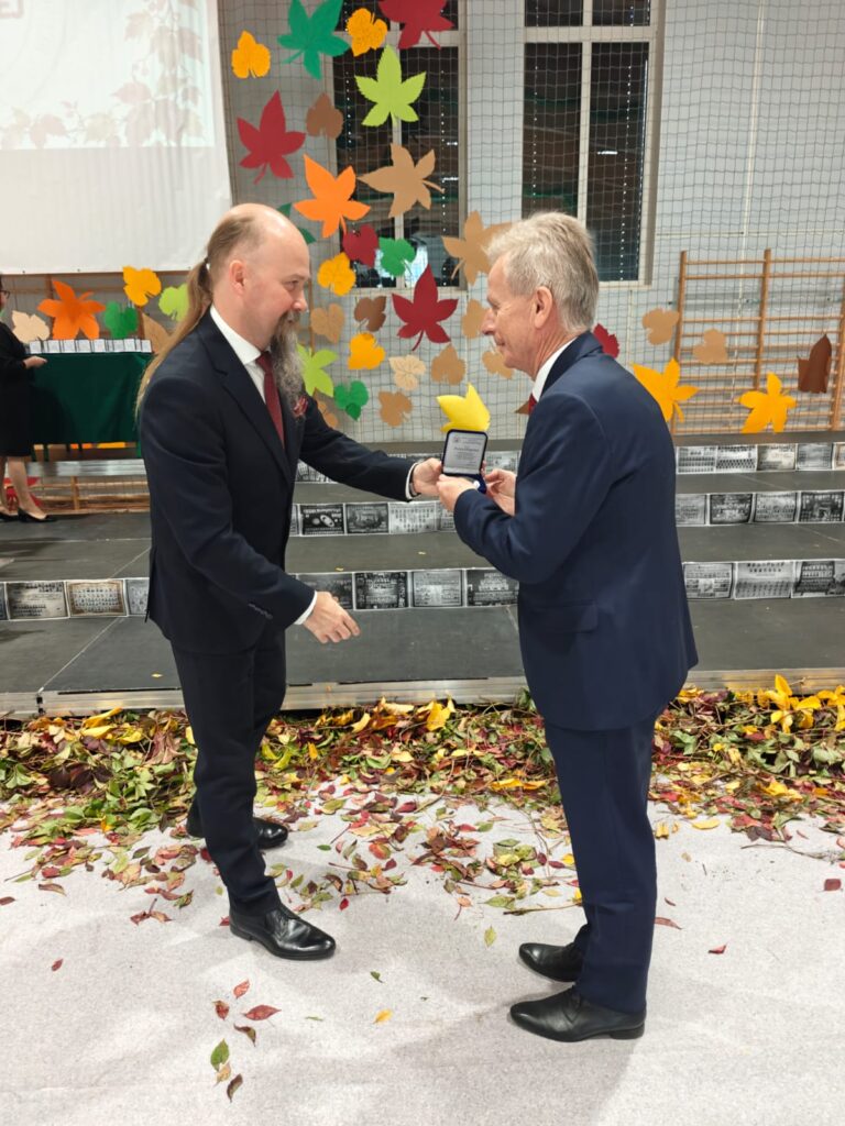 Dwaj mężczyźni w garniturach wymieniają uścisk dłoni podczas uroczystości szkolnej.