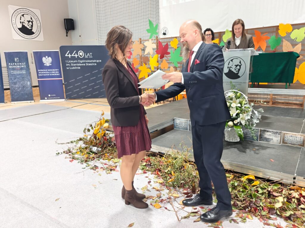 Mężczyzna wręcza dokument kobiecie w czerwonej sukience.