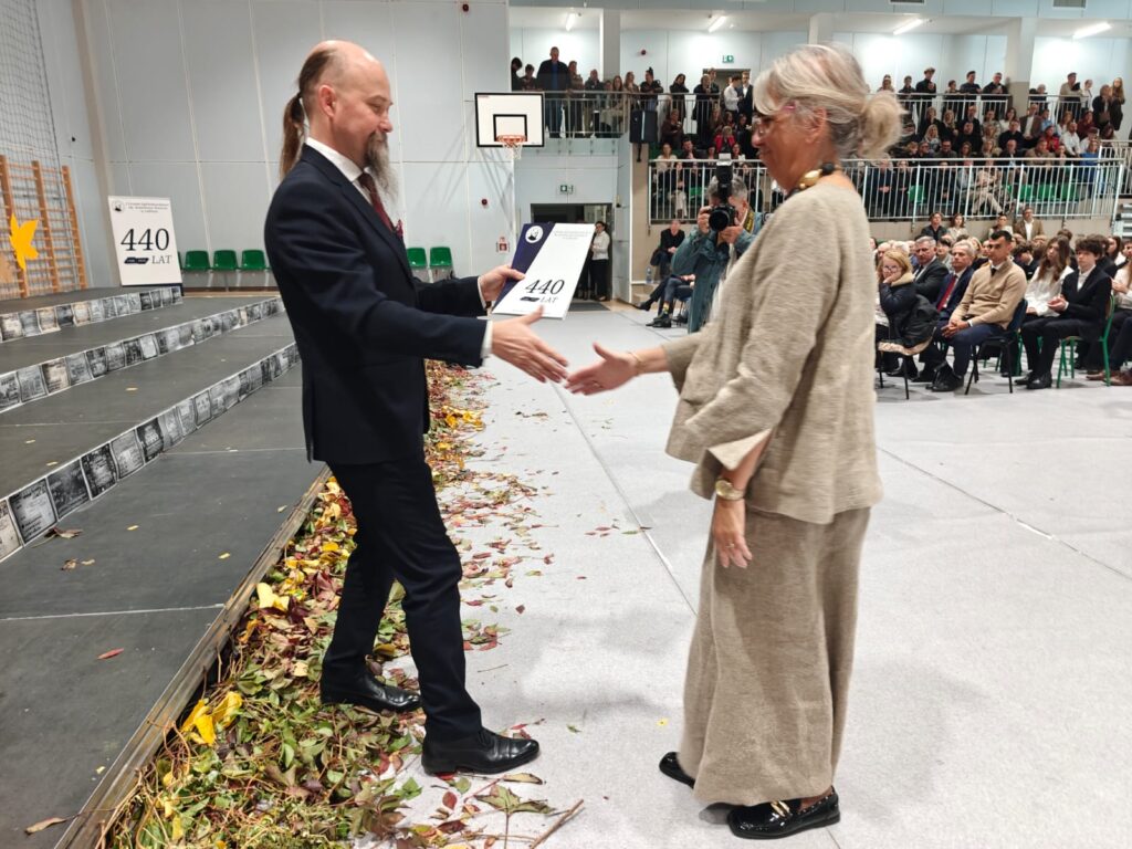 Mężczyzna wręcza wyróżnienie starszej kobiecie w jasnym stroju.