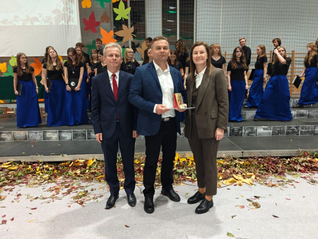 Trzy osoby pozują do zdjęcia z pamiątkowym wyróżnieniem, w tle stoi chór.