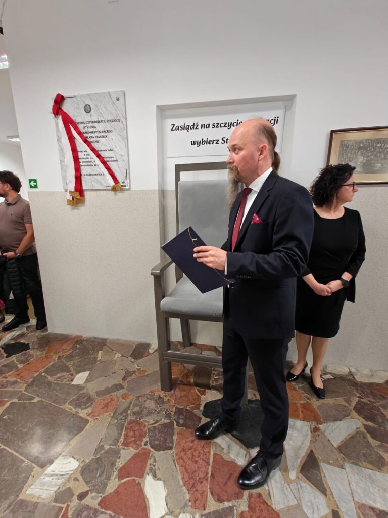 Mężczyzna w granatowym garniturze przemawia przy odsłoniętej tablicy pamiątkowej, trzymając teczkę z dokumentami. Obok stoi kobieta w czarnej sukience.